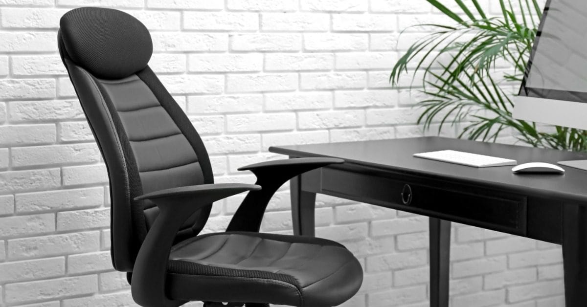 Black executive office chair with padded headrest and armrests beside a black desk, monitor, and green plant.
