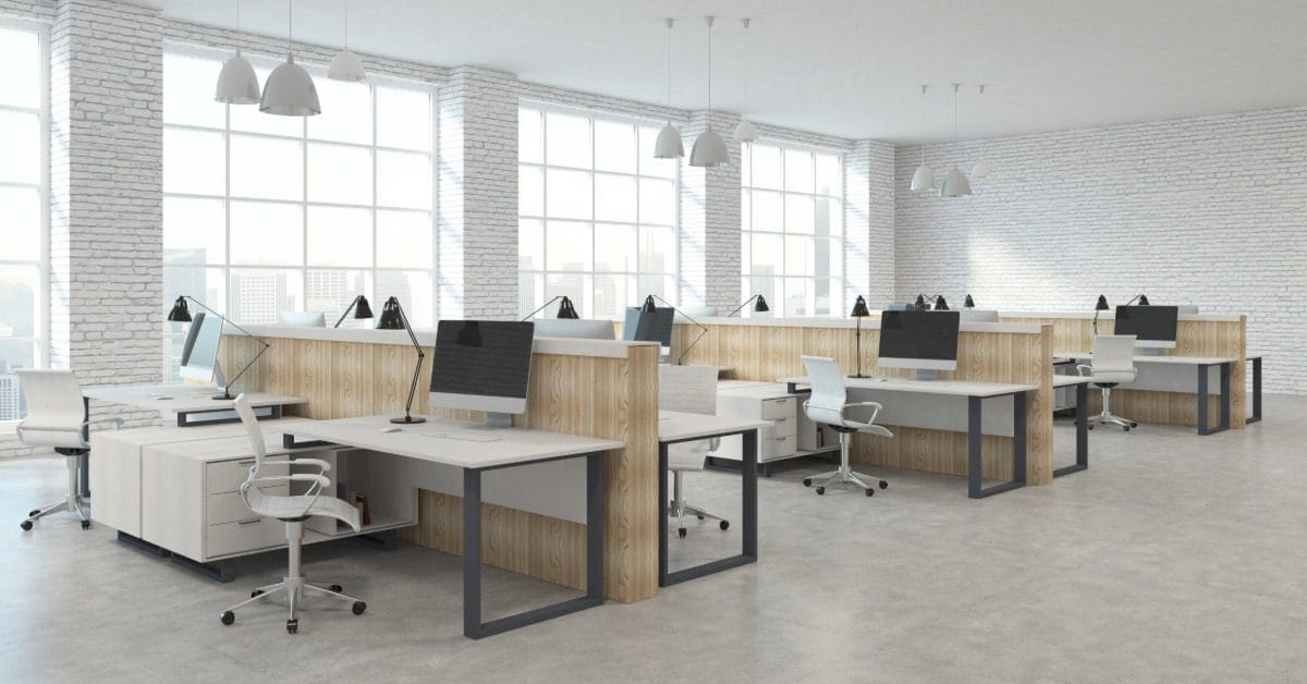 A modern open office with white desks, wood partitions, desktop computers, rolling chairs, and large sunlit windows.
