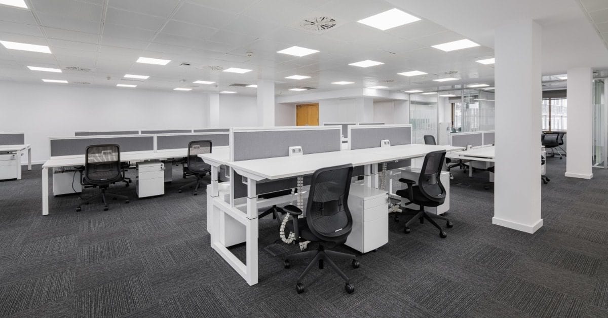 A modern open office with white desks, gray cubicles, black ergonomic chairs, and bright overhead lighting.