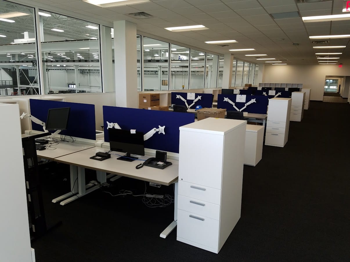 Friant benching workstations with blue privacy screens and monitor arms along the plant windows at United Grinding North America in Miamisburg, Ohio