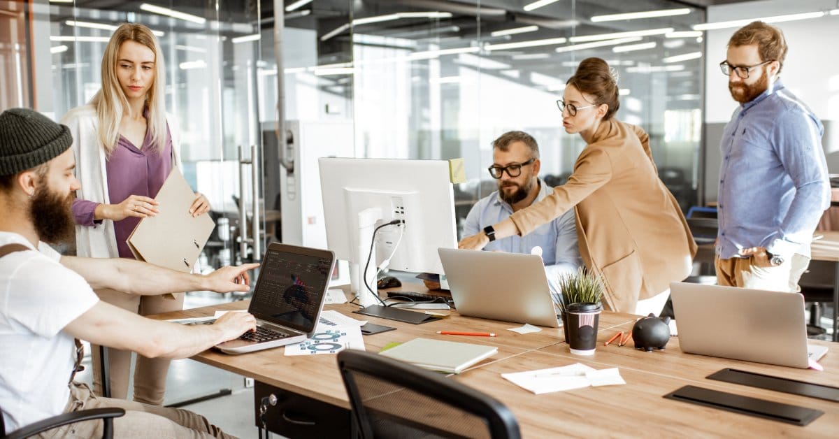 Coworkers collaborating at a shared wooden desk in a modern hybrid office.