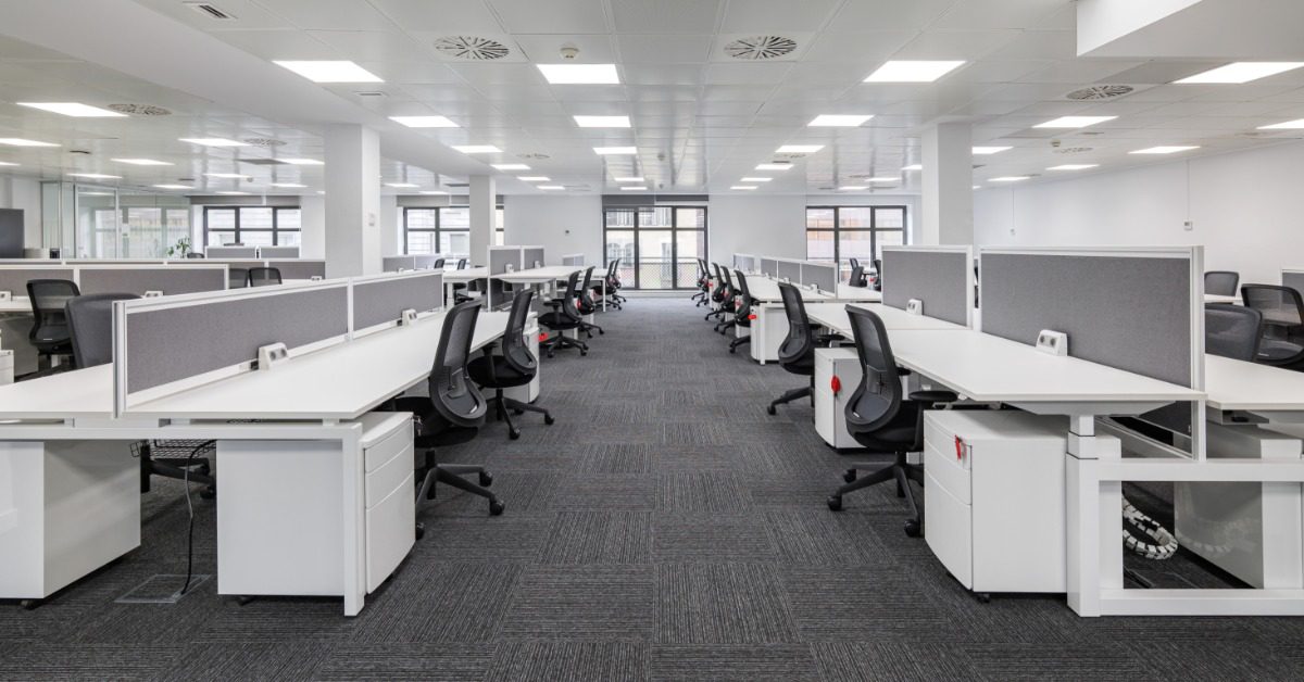 A spacious open office with rows of white desks, gray privacy panels, black task chairs, and bright overhead lighting.