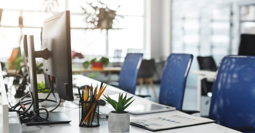 A bright open office with computer monitors, blue task chairs, a white desk, pencils, documents, and a small plant.