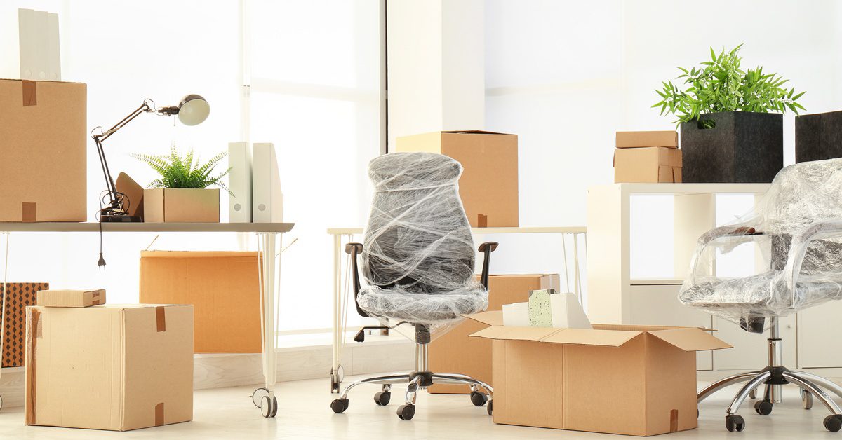 Office furniture wrapped for moving sits among packed boxes in a bright workspace with desks, chairs, and plants.