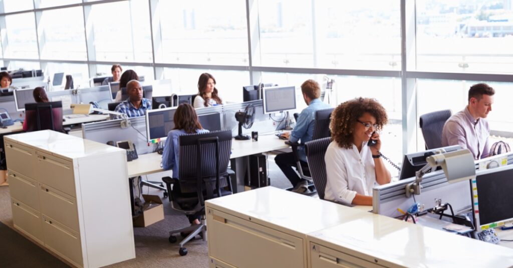 An open-plan office with bright floor-to-ceiling windows and employees working at shared desk spaces.