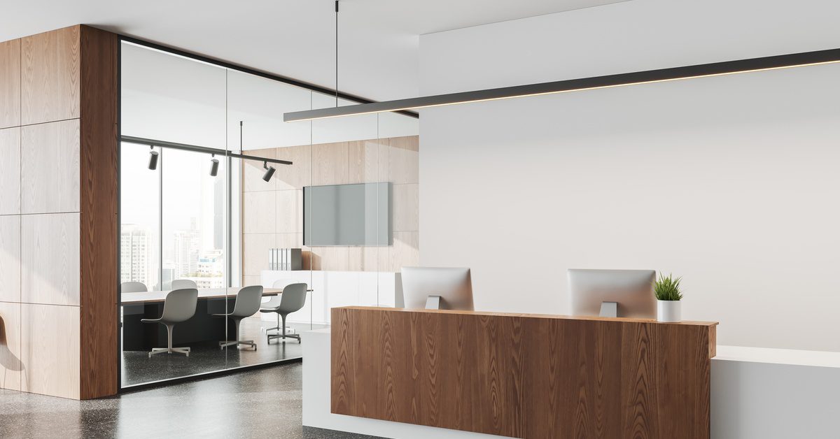 A modern office reception with a walnut front desk, dual monitors, glass meeting room, and bright open lobby.