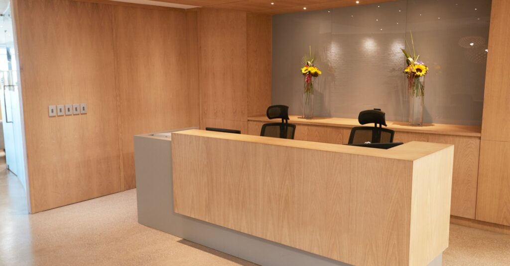 A light wood reception desk with two workstations, floral displays, and a warm, modern office lobby interior.