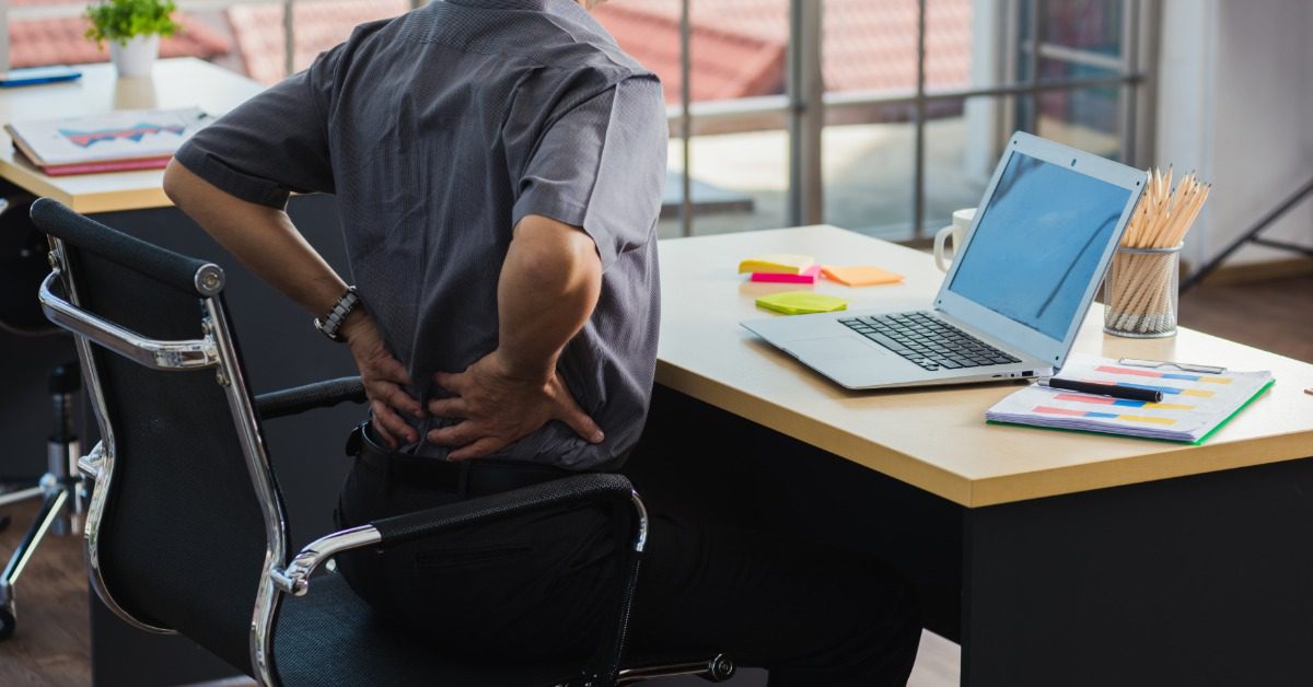 Office worker holds his lower back while seated at a desk with a laptop, showing discomfort from prolonged sitting.