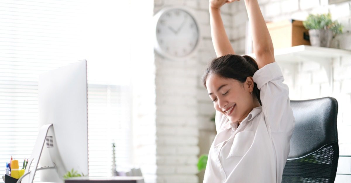 Office worker stretches overhead while seated at a desk with a computer in a bright workspace to relieve muscle tension.